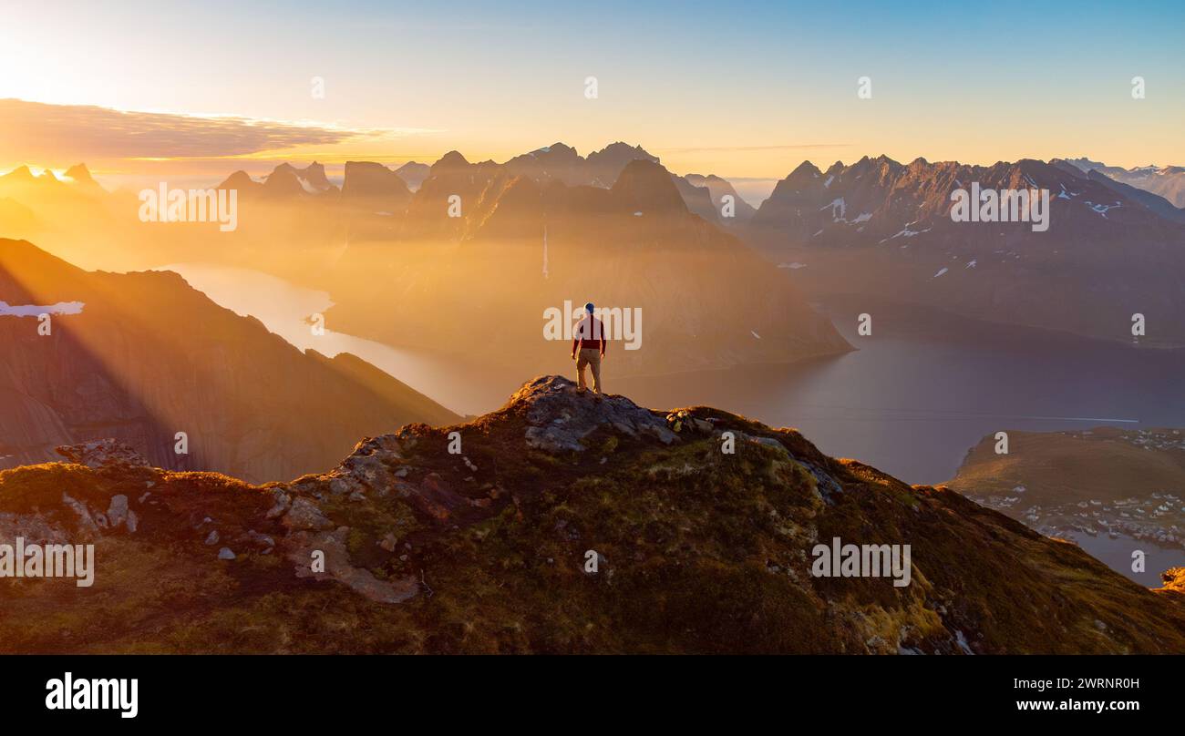 Male hiker enjoying mazing midnight sun on top of mountain in Reine, Lofoten, Norway Stock Photo