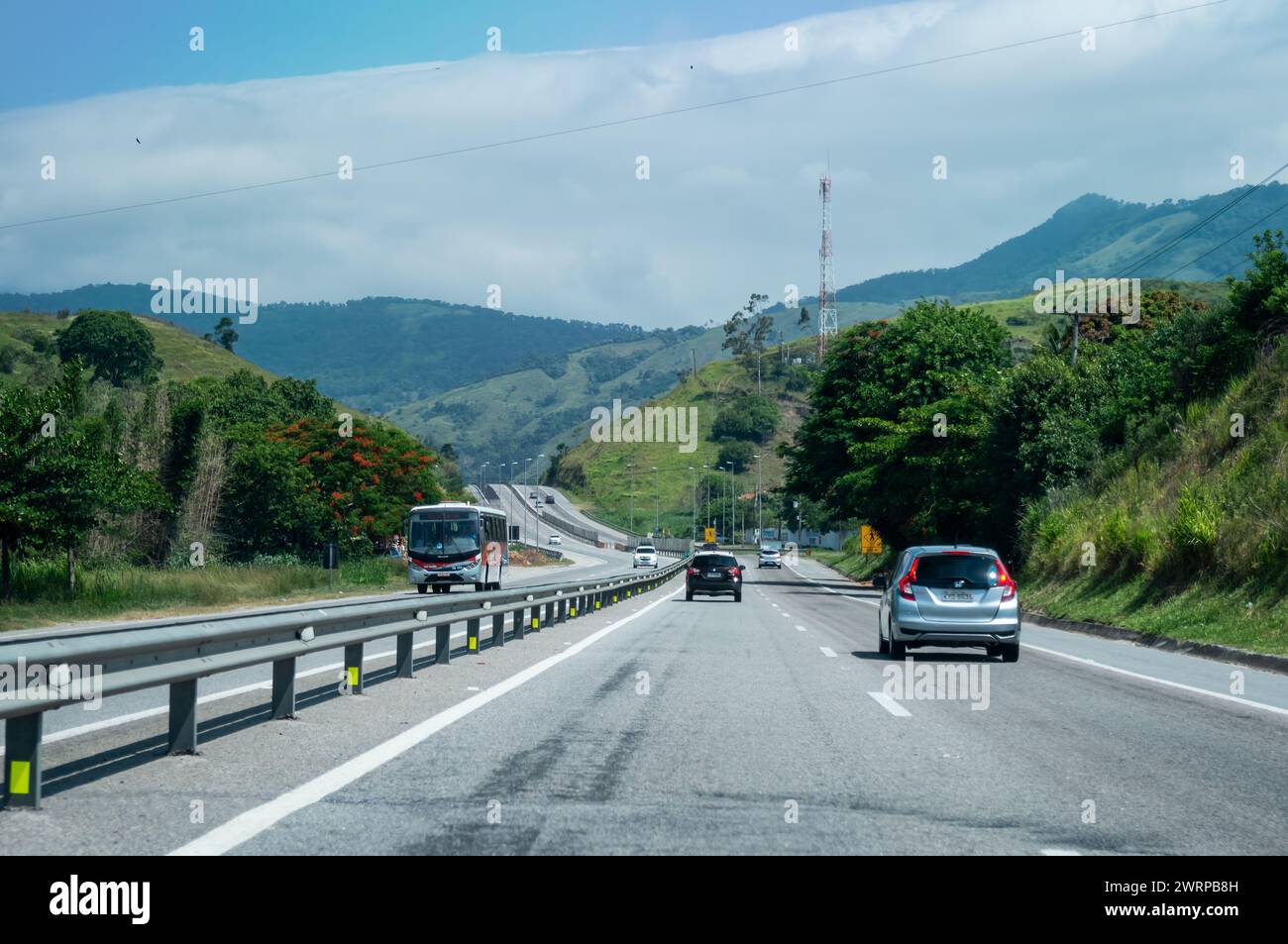 View at KM 9 of Via Lagos highway with cars passing by with a big mountain covered by Atlantic forest vegetation at back under summer clear blue sky. Stock Photo