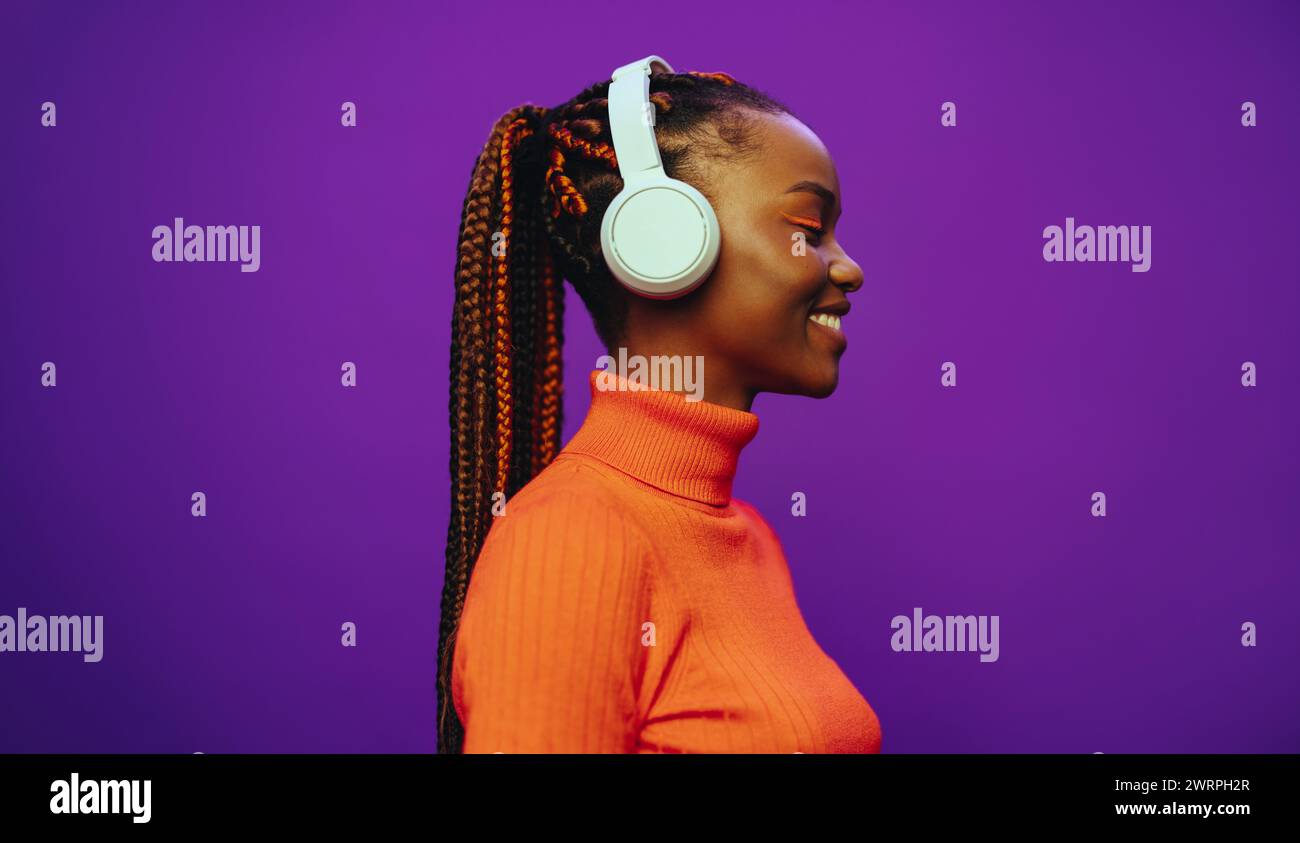 Smiling young woman with purple two-tone braids and trendy makeup stands in a studio, wearing casual clothing. She listens to music with headphones, e Stock Photo