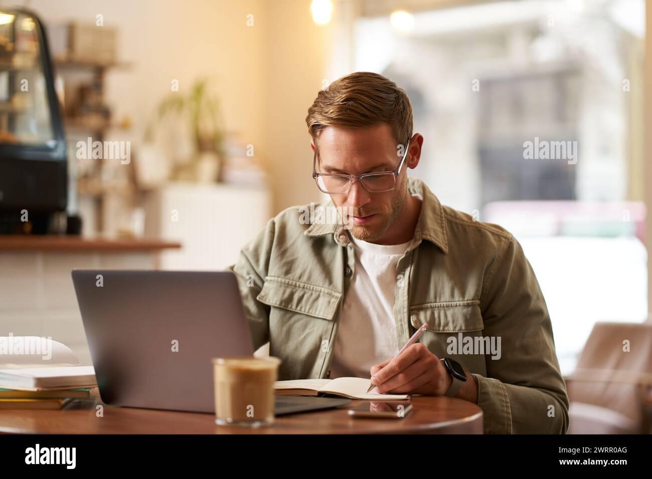 Image of focused young man in glasses, sitting in cafe, making notes, studying, attending online course, learning on remote from quite coffee shop Stock Photo