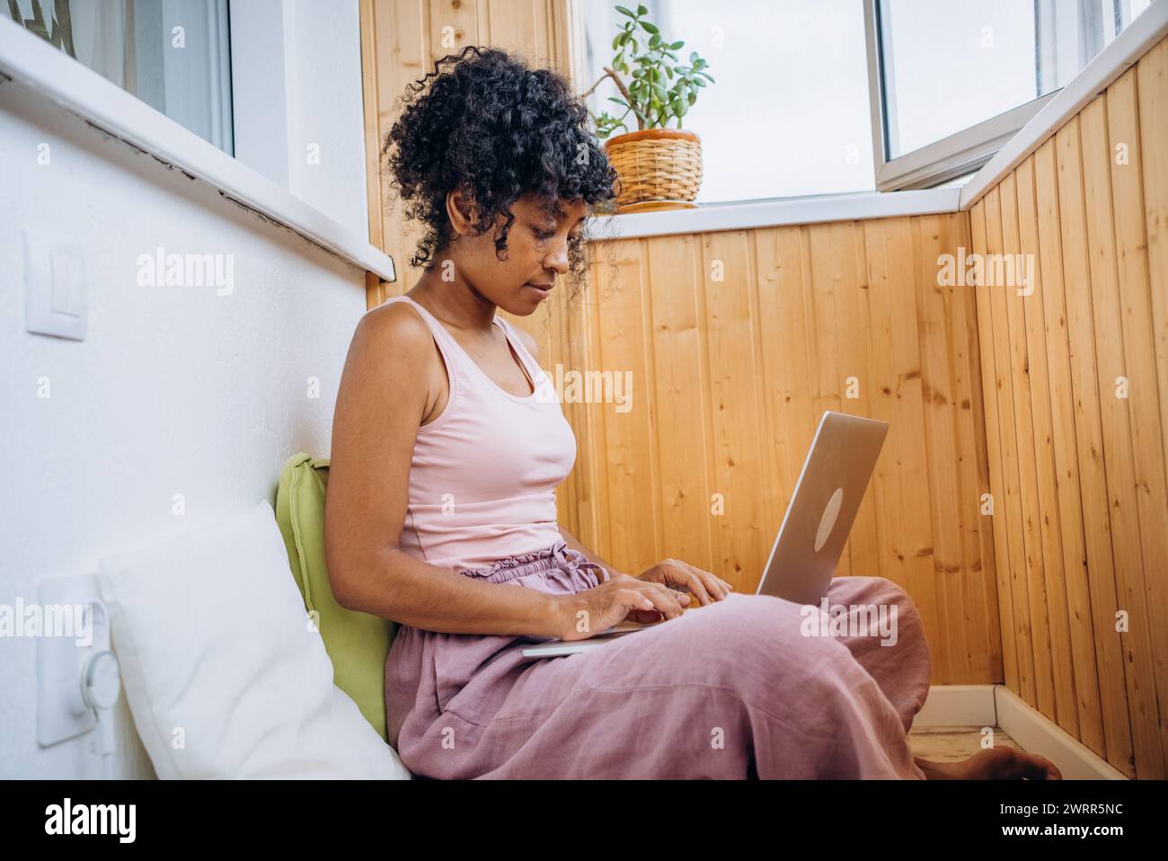 A young woman in casual attire focuses intently on her laptop in a cozy ...