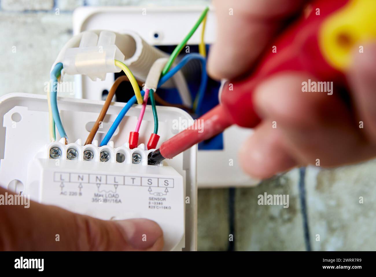 Electrician connecting colored wires to terminals of wall mounted thermostat for floor heating in residential building during renovation. Stock Photo