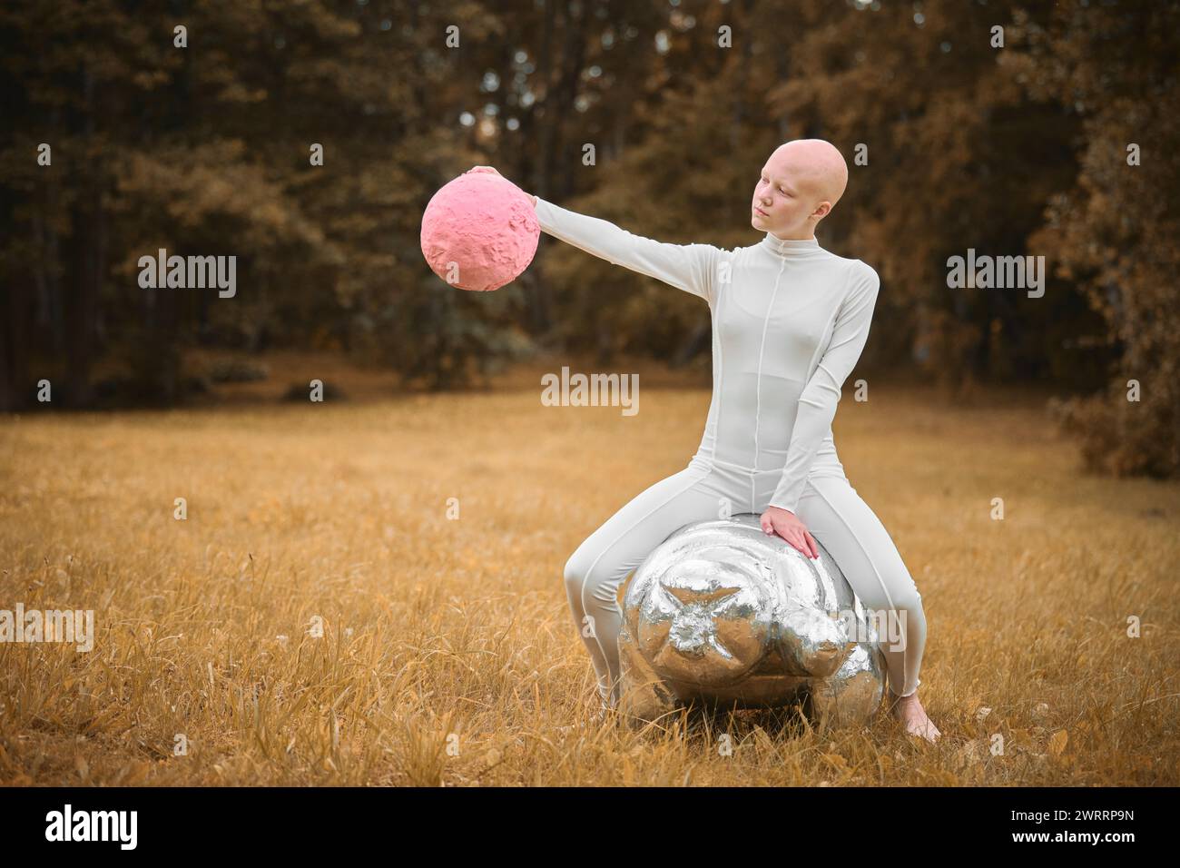 Young hairless girl with alopecia in white cloth sits on tardigrade figure and holds pink ball in hand on fall lawn park, surreal scene with bald teen Stock Photo