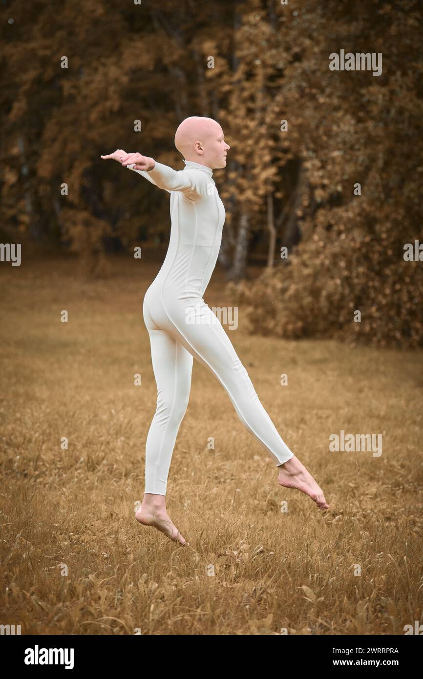 Full length portrait of young hairless girl ballerina with alopecia in tight white suit prepares to jump on fall lawn in park, symbolizing overcoming Stock Photo