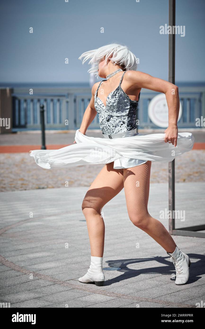Young sexy girl in space silver micro skirt dancing with white silk scarf waving gracefully, female outdoor dance performance on seaside promenade cre Stock Photo