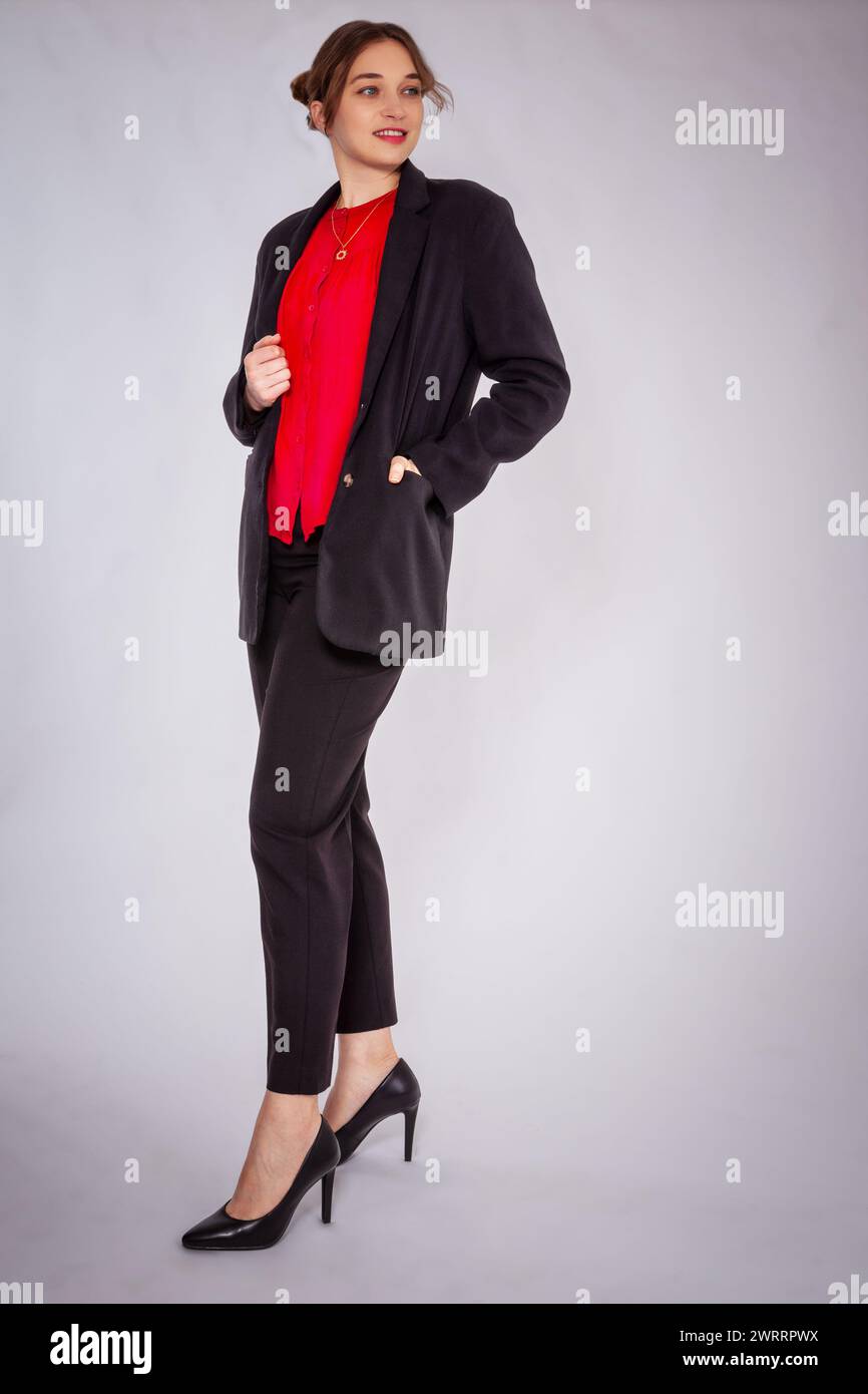 A young, blonde woman with a bun wears a black trouser suit and a red blouse. She combines this with elegant high heels. She poses confidently in fron Stock Photo