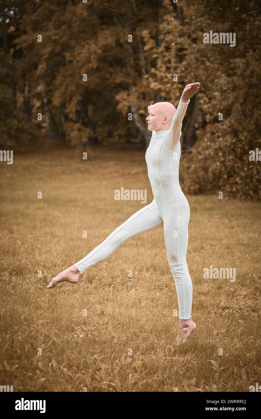 Full length portrait of young hairless girl ballerina with alopecia in tight white suit prepares to jump on fall lawn in park, symbolizing overcoming Stock Photo