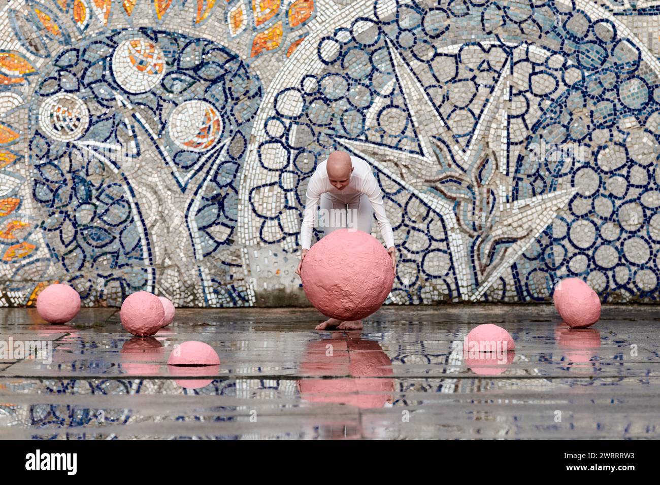 Young hairless girl with alopecia in white futuristic suit dancing outdoor smoothly and takes pink ball on abstract mosaic Soviet background, symboliz Stock Photo