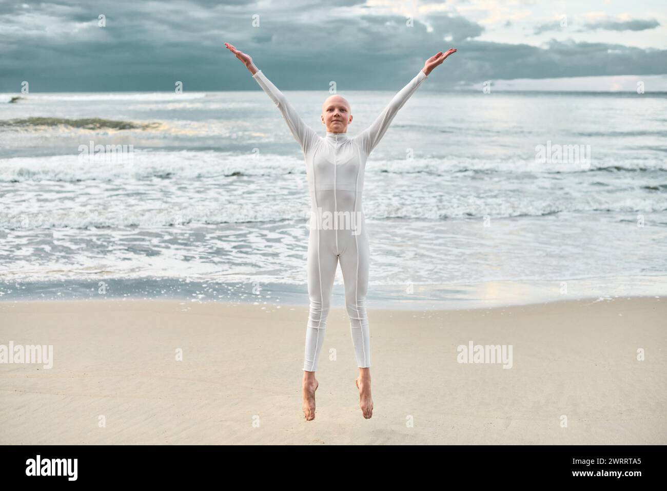 Joyful hairless girl with alopecia in white futuristic suit jumping spreading arms and legs apart on sea sandy beach, metaphoric performance with bald Stock Photo