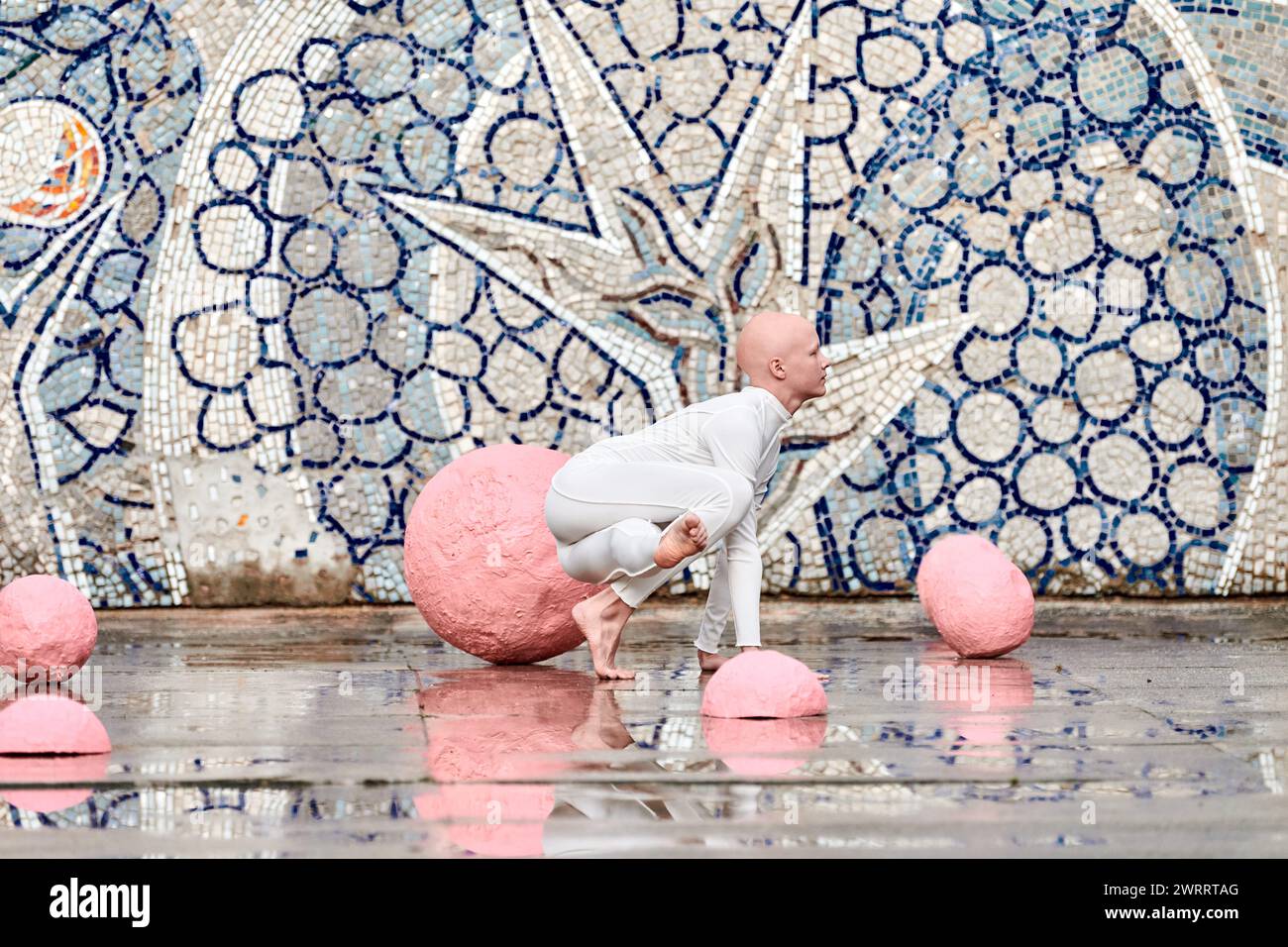 Young hairless girl ballerina with alopecia in white futuristic suit dancing outdoor among pink spheres on abstract mosaic Soviet background, symboliz Stock Photo