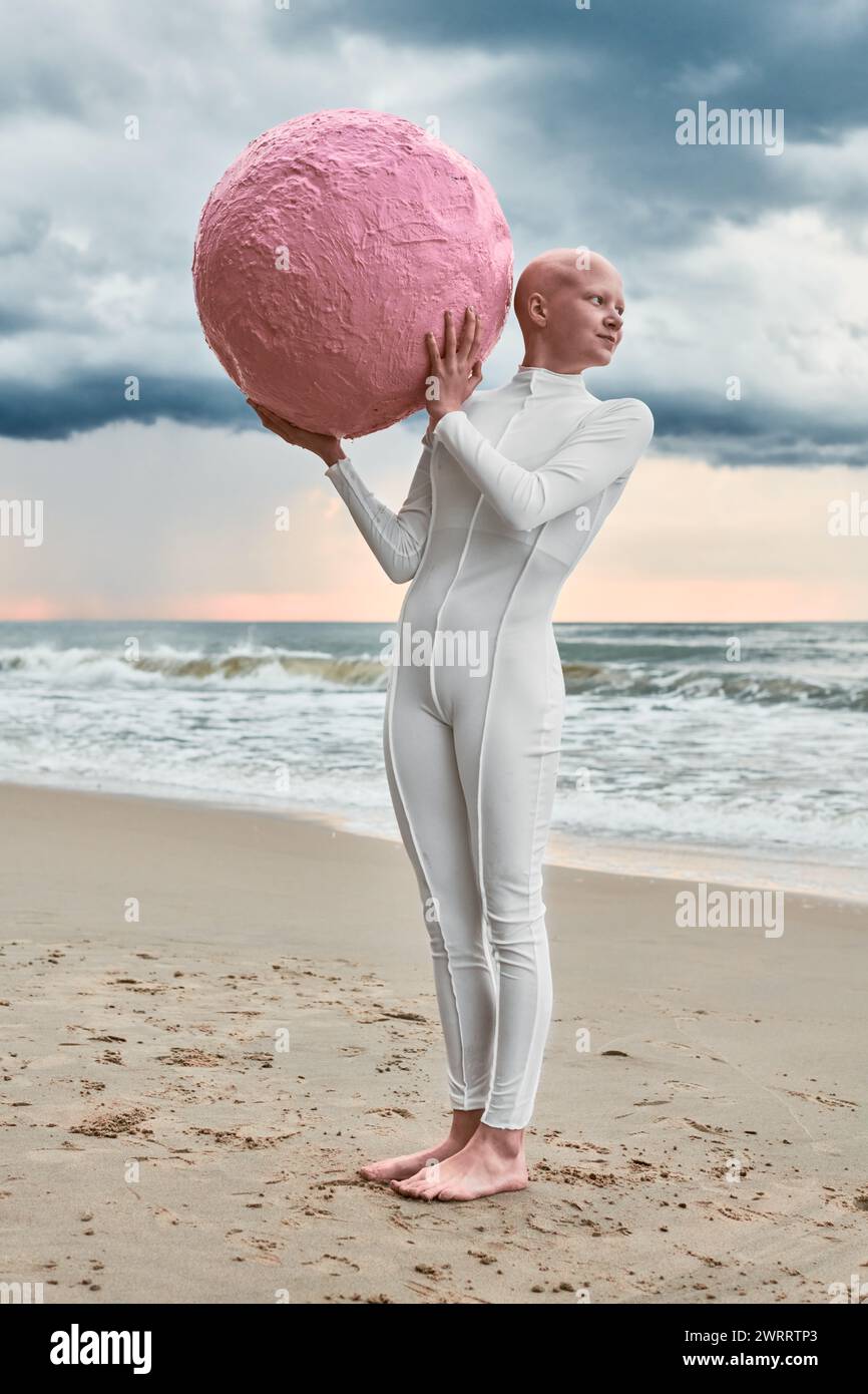 Full length portrait of hairless girl with alopecia in white futuristic suit standing on stone sea beach and hold pink sphere, metaphoric surreal perf Stock Photo