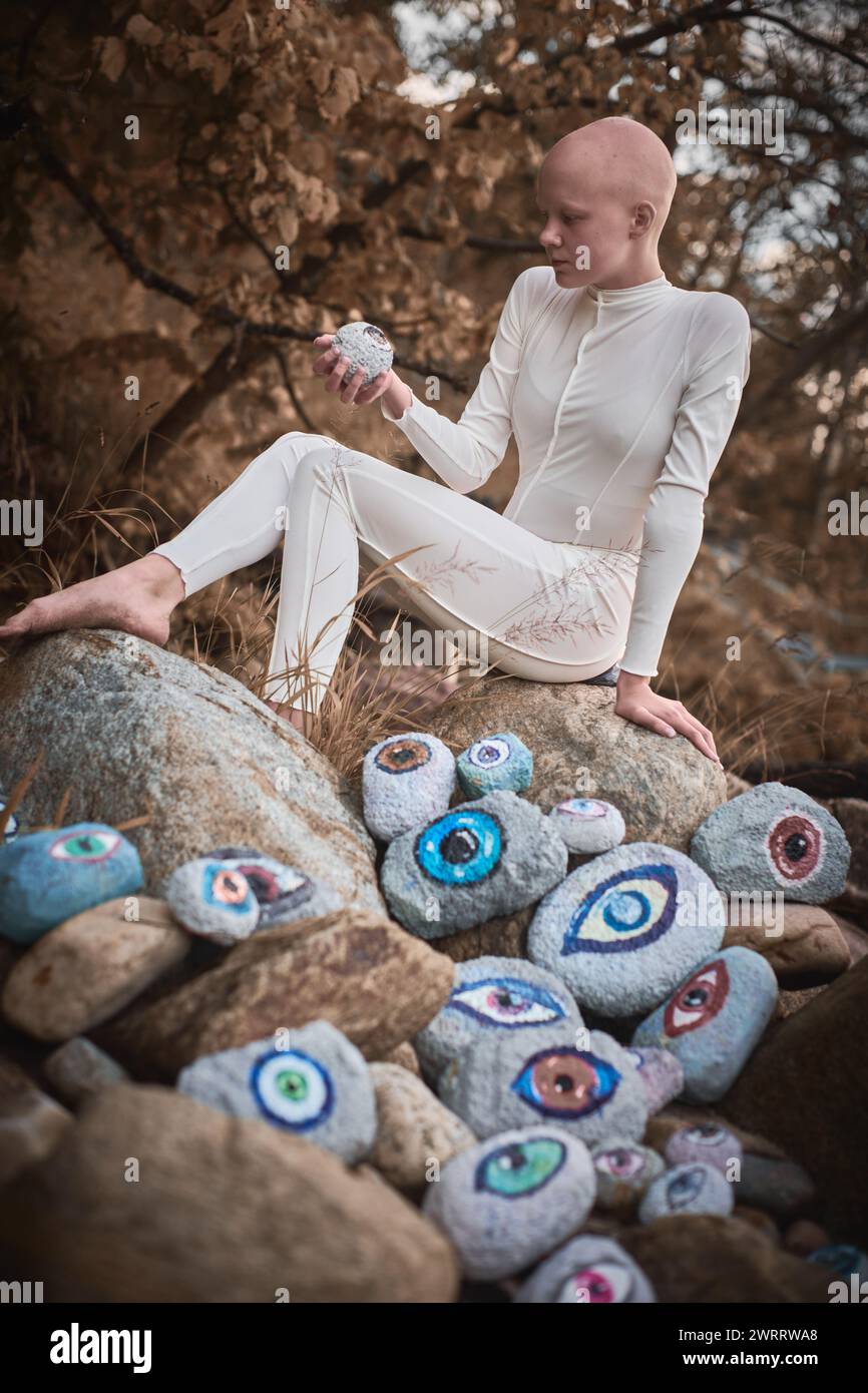 Young hairless girl with alopecia in white futuristic costume pensively examines stone with eye at surreal landscape, performance symbolizes introspec Stock Photo