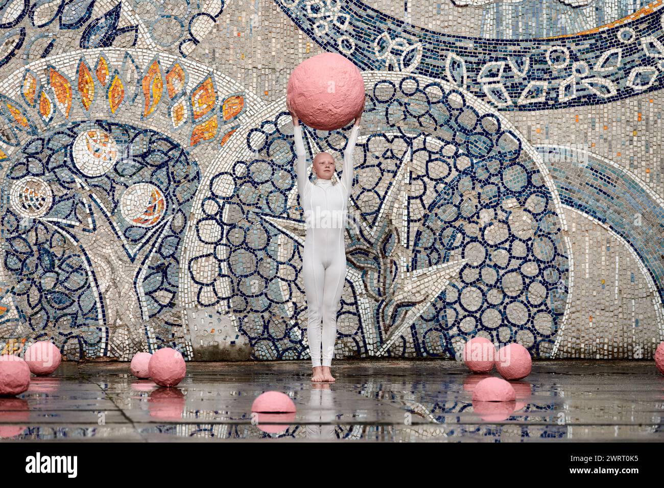 Young hairless girl with alopecia in white futuristic suit dancing outdoor smoothly holding pink ball on abstract mosaic Soviet background, symbolizes Stock Photo