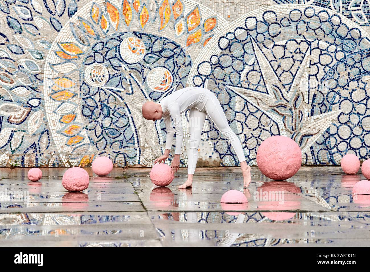 Young hairless girl with alopecia in white futuristic suit dancing outdoor smoothly and takes pink ball on abstract mosaic Soviet background, symboliz Stock Photo