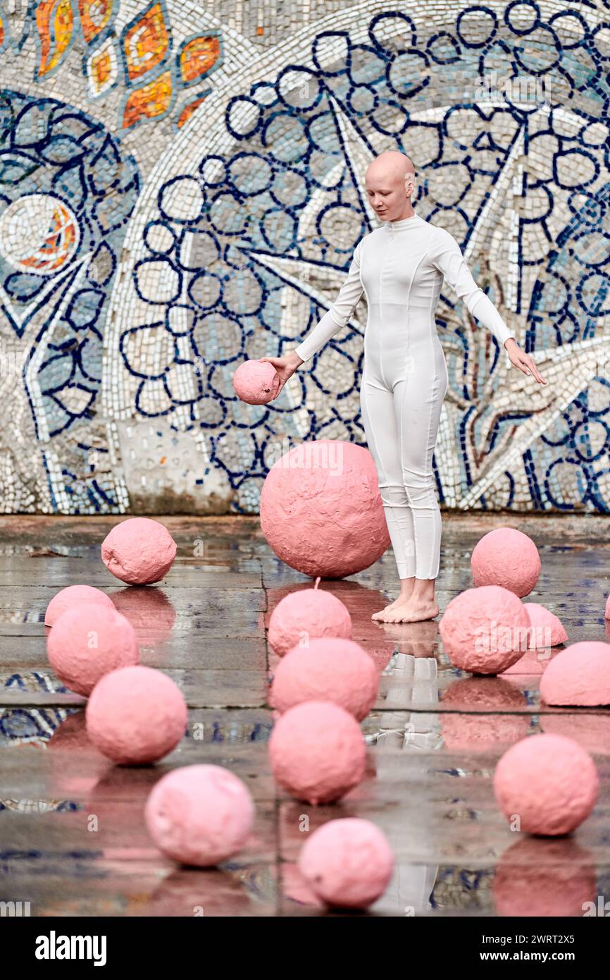 Young hairless girl ballerina with alopecia in white futuristic suit dancing outdoor among pink spheres on abstract mosaic Soviet background, symboliz Stock Photo