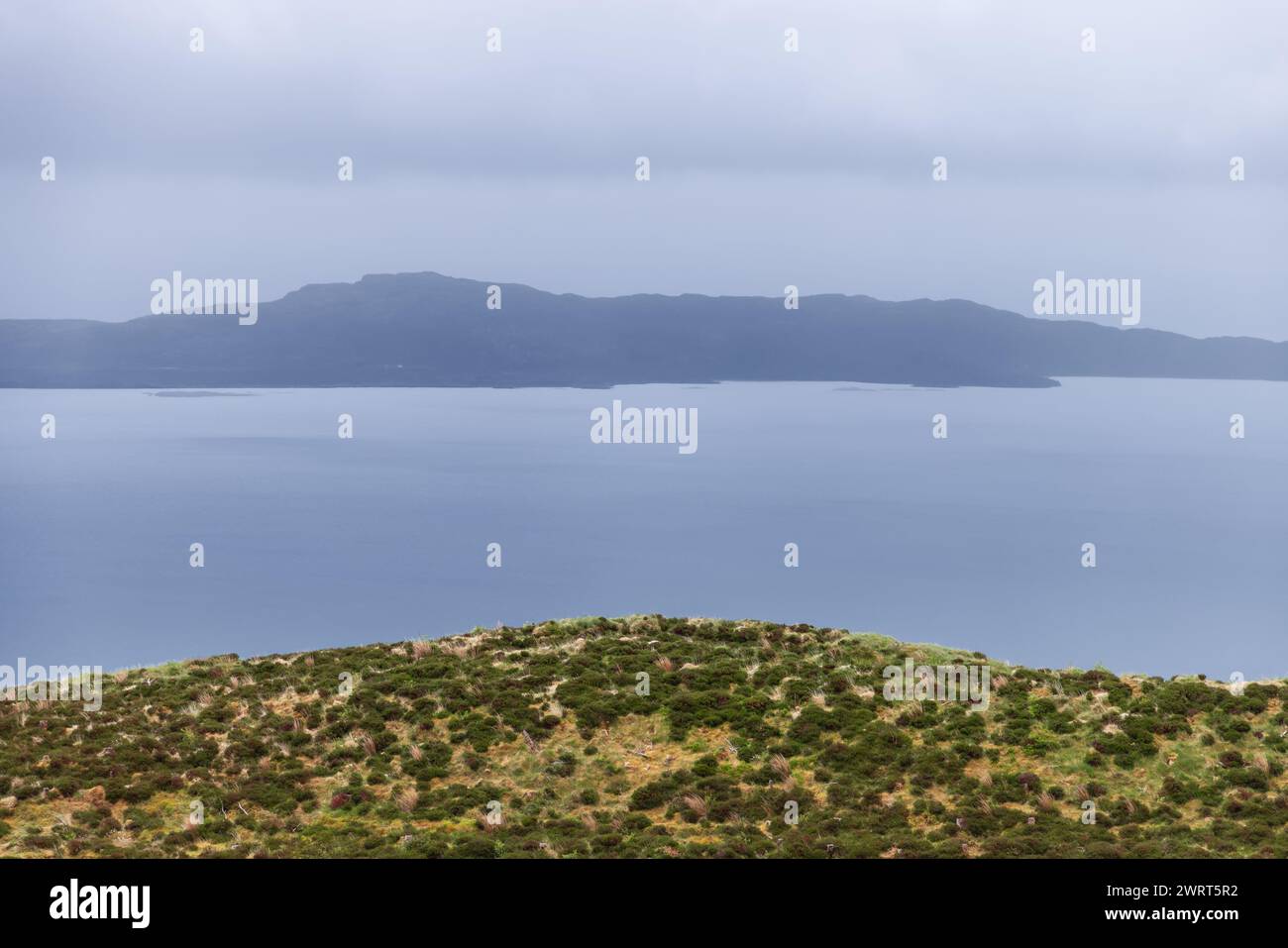A serene expanse of sea blurs with the hazy outline of Raasay Island under a soft grey sky, framed by the wild heath of the Isle of Skye Stock Photo