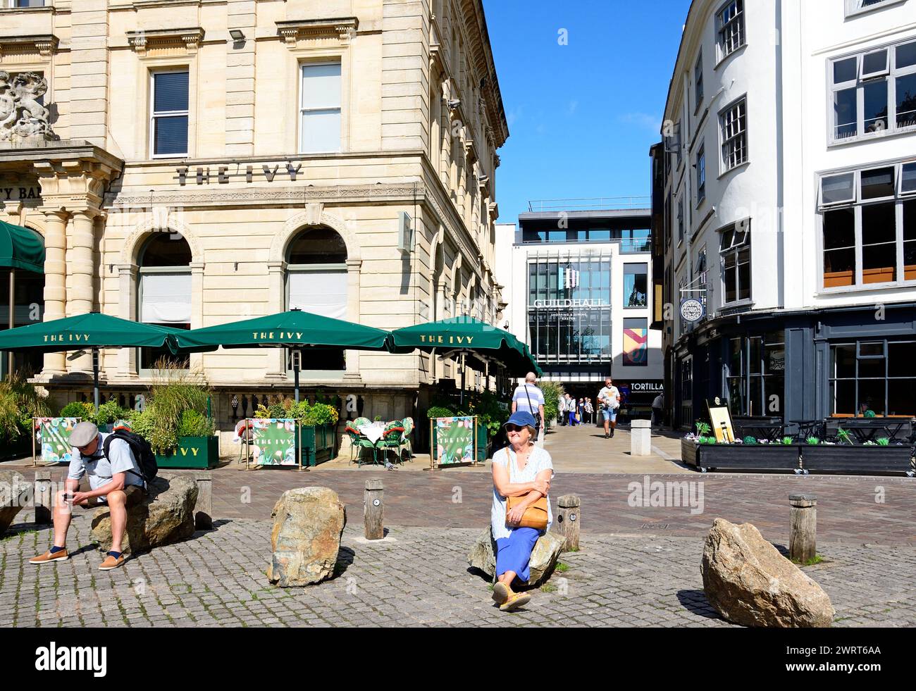 The Ivy Bistro and side street along Cathedral Yard with tourists sitting on rocks in the foreground, Exeter, Devon, UK, Europe. Stock Photo