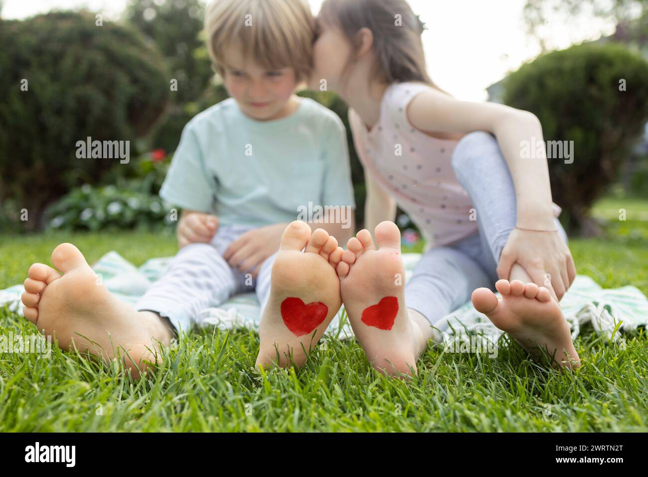 two barefoot children. a boy and a girl, 7-8 years old, sitting on the grass, red hearts drawn on their feet. as a symbol of love. children's tender f Stock Photo
