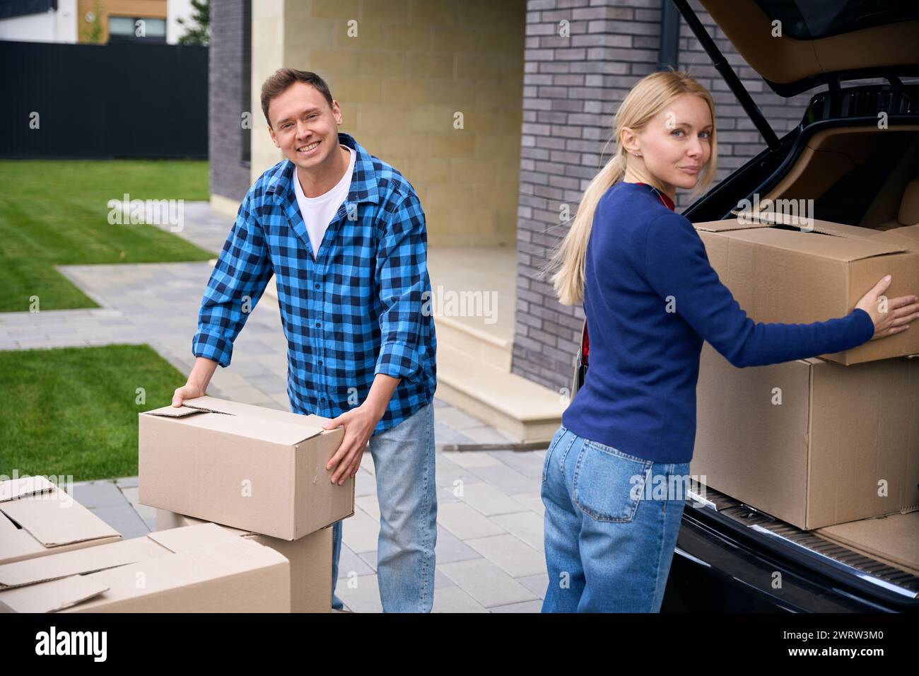 Contented couple standing near car and loading its trunk with cardboard boxes during house move Stock Photo