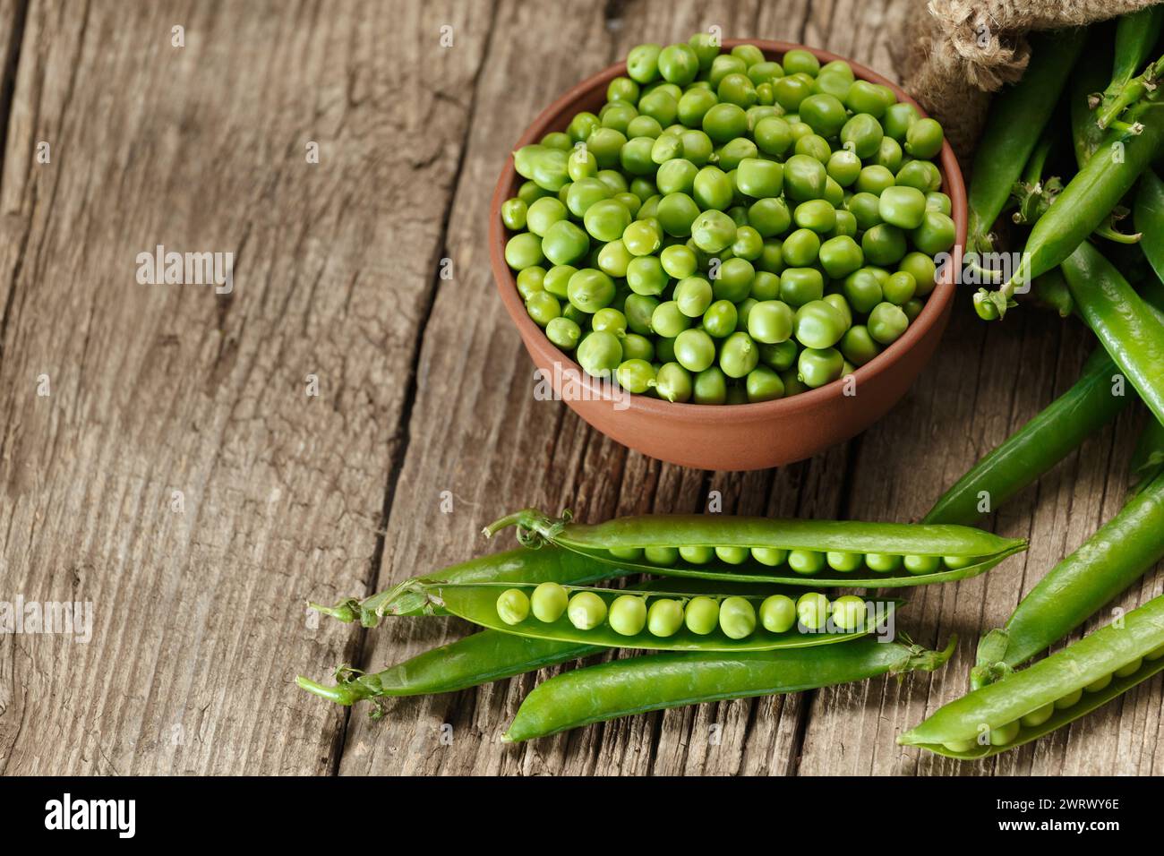 Pods of fresh green peas in a burlap bag, shelled peas in a clay bowl, sweet organic green peas in closed and open pods on an aged wooden background. Stock Photo
