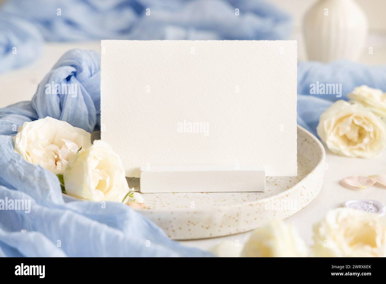 Blank card near light blue tulle fabric and cream flowers on plate close up, copy space. Wedding stationery mockup. Romantic table place with horizont Stock Photo