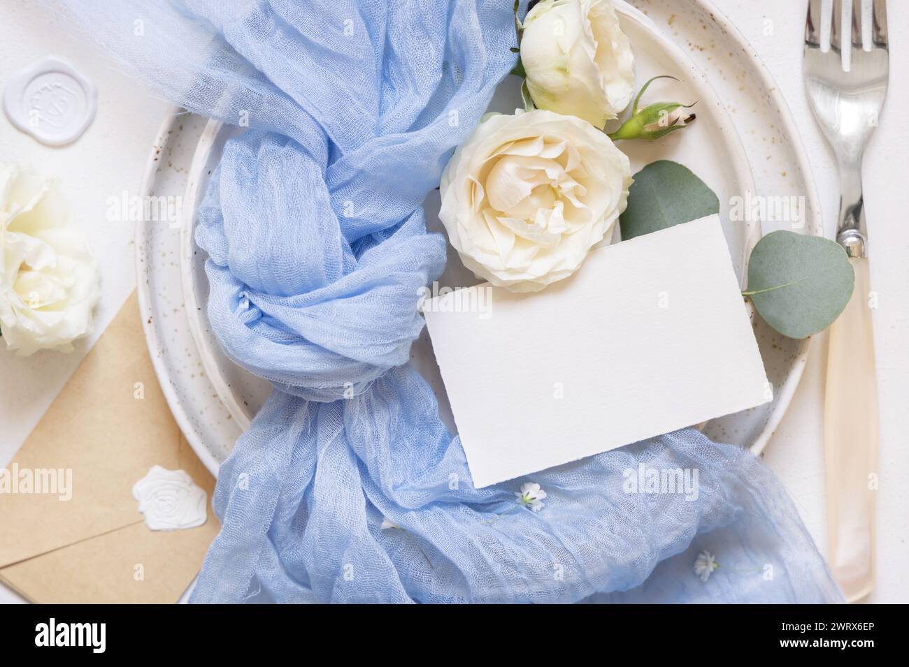 Blank card near light blue tulle fabric and cream roses on plates top view, copy space. Wedding stationery mockup. Romantic table place with horizonta Stock Photo