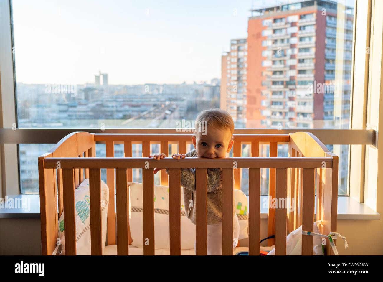 The baby stands in the crib and looks bored inside the room, not paying