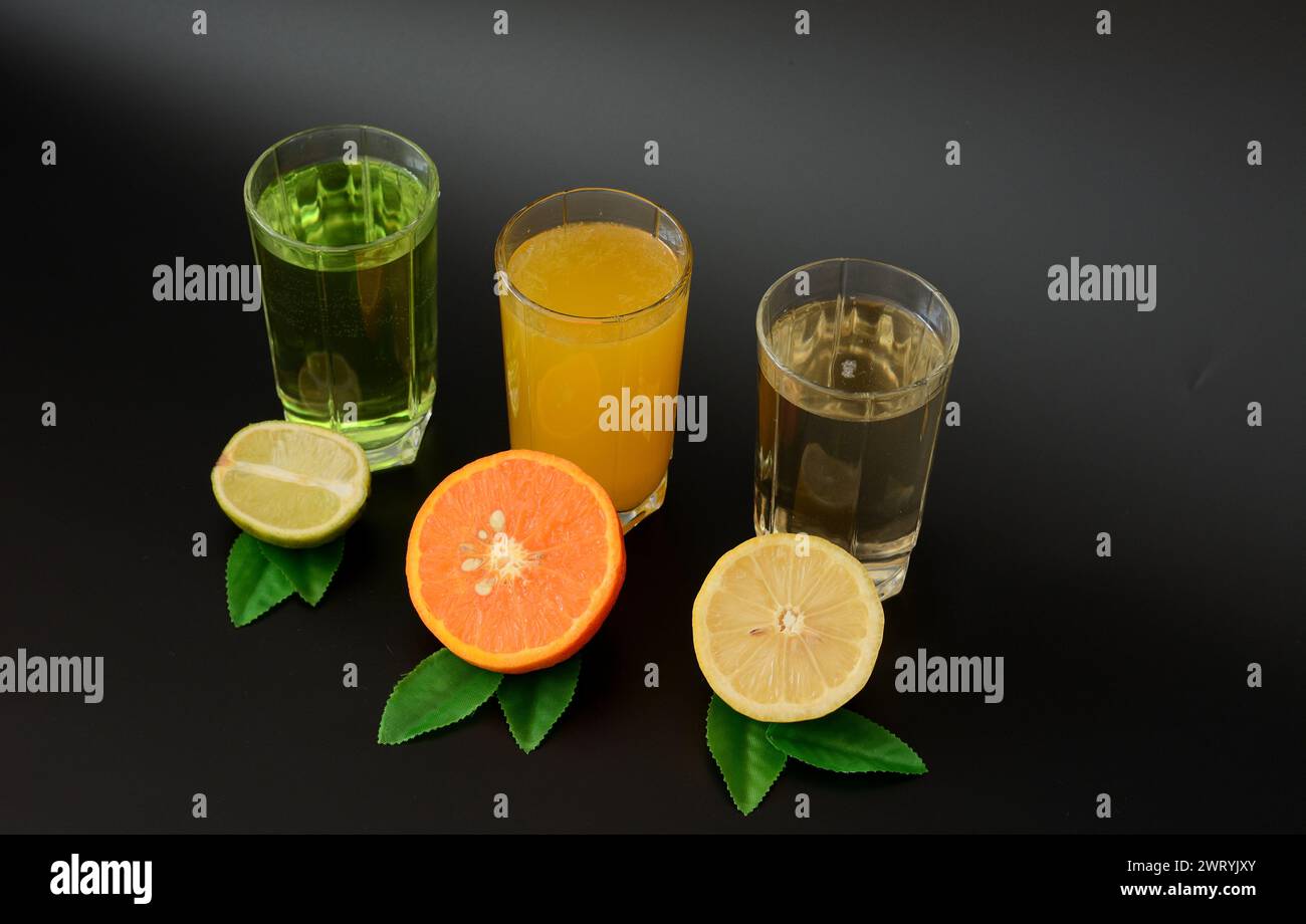 Three glasses with different citrus juices stand in a row on a black background, next to the halves of a lemon, orange and lime with leaves. Close-up. Stock Photo