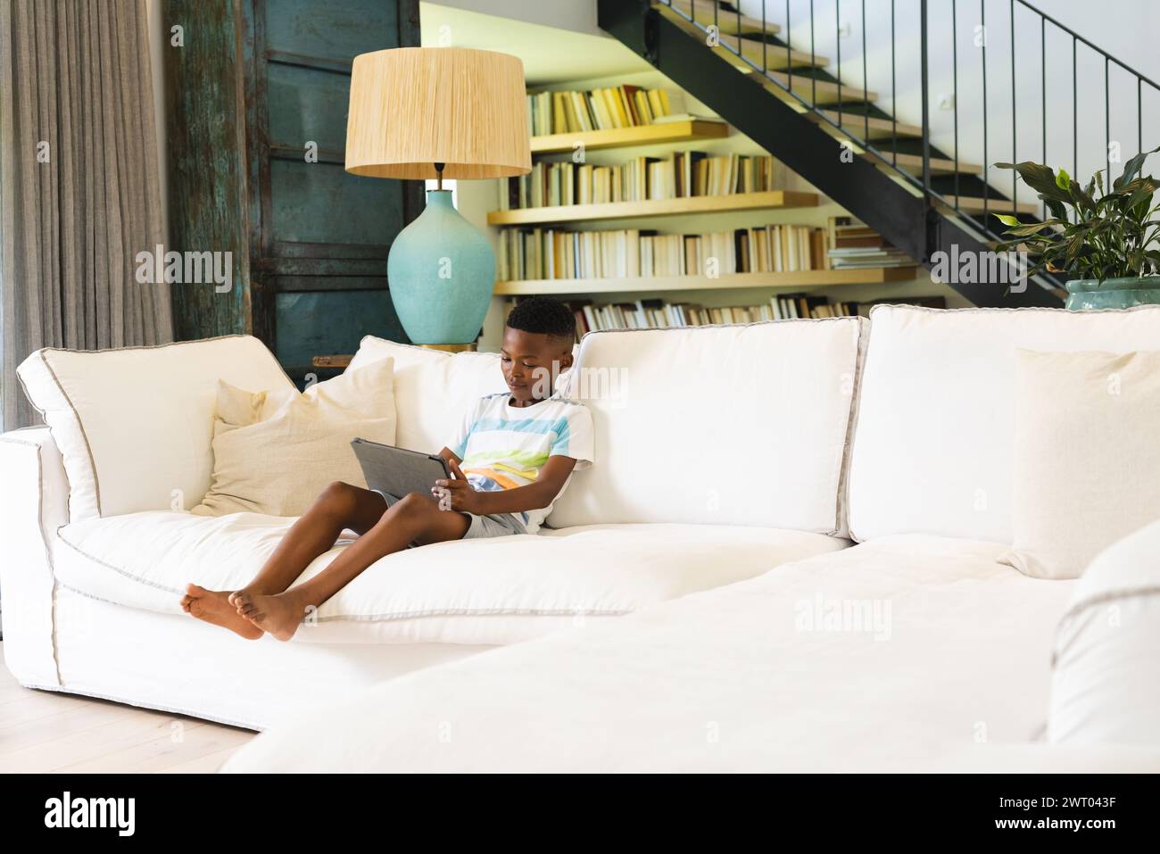 A young African American boy is engrossed in a tablet on a white sofa with copy space at home Stock Photo