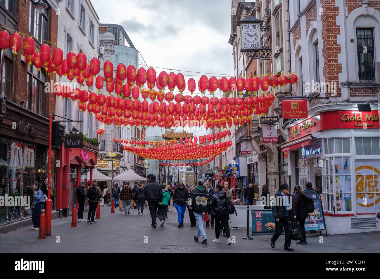 People dressed warmly walk on busy vibrant Wardour street in Chinatown London. Strings of traditional Chinese red lanterns stretch across the street. Stock Photo