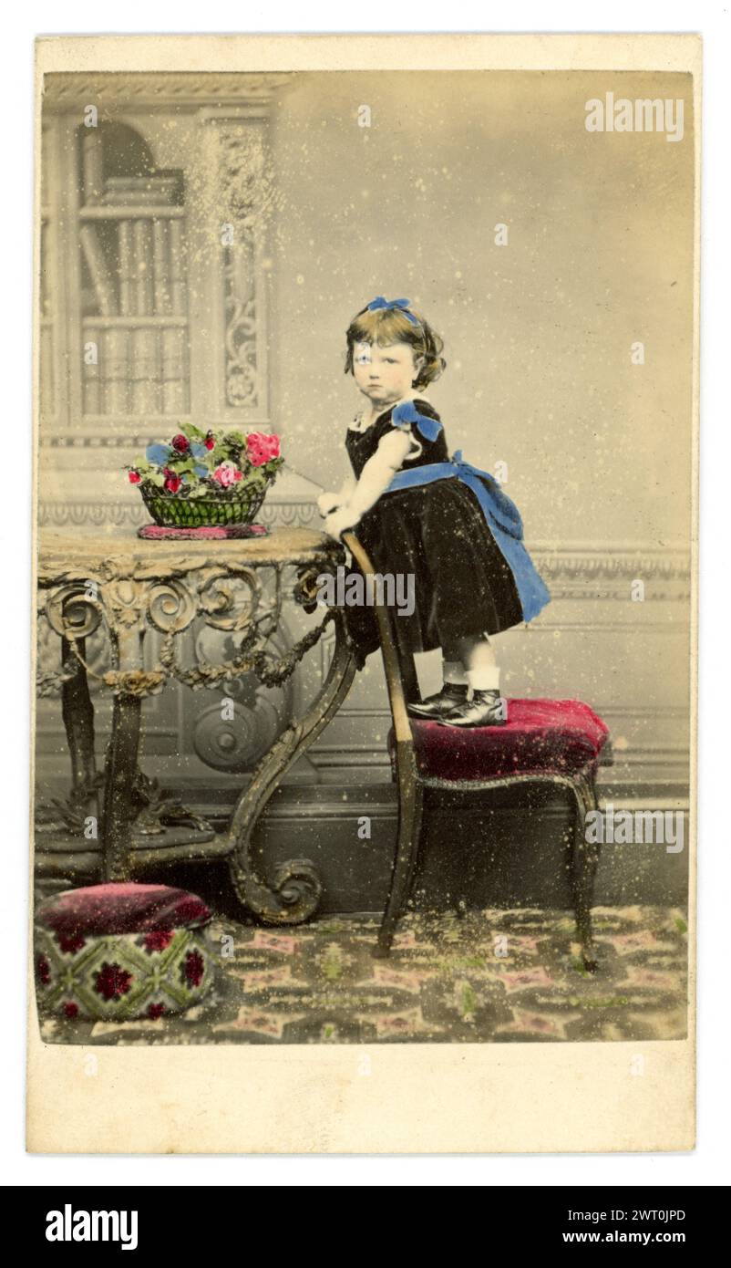 Original, charming, tinted Carte de Visite (visiting card or CDV)  of cute little Victorian girl, Victorian child, aged about 2 or 3 years old, wearing a black velvet dress, with blue triim and ribbon and a blue hairband, looking serious, standing on a velvet stool, studio of Holloway School of Photography, G Williams, Pear Tree cottage, Holloway Rd. London, U.K. Circa 1860's Stock Photo