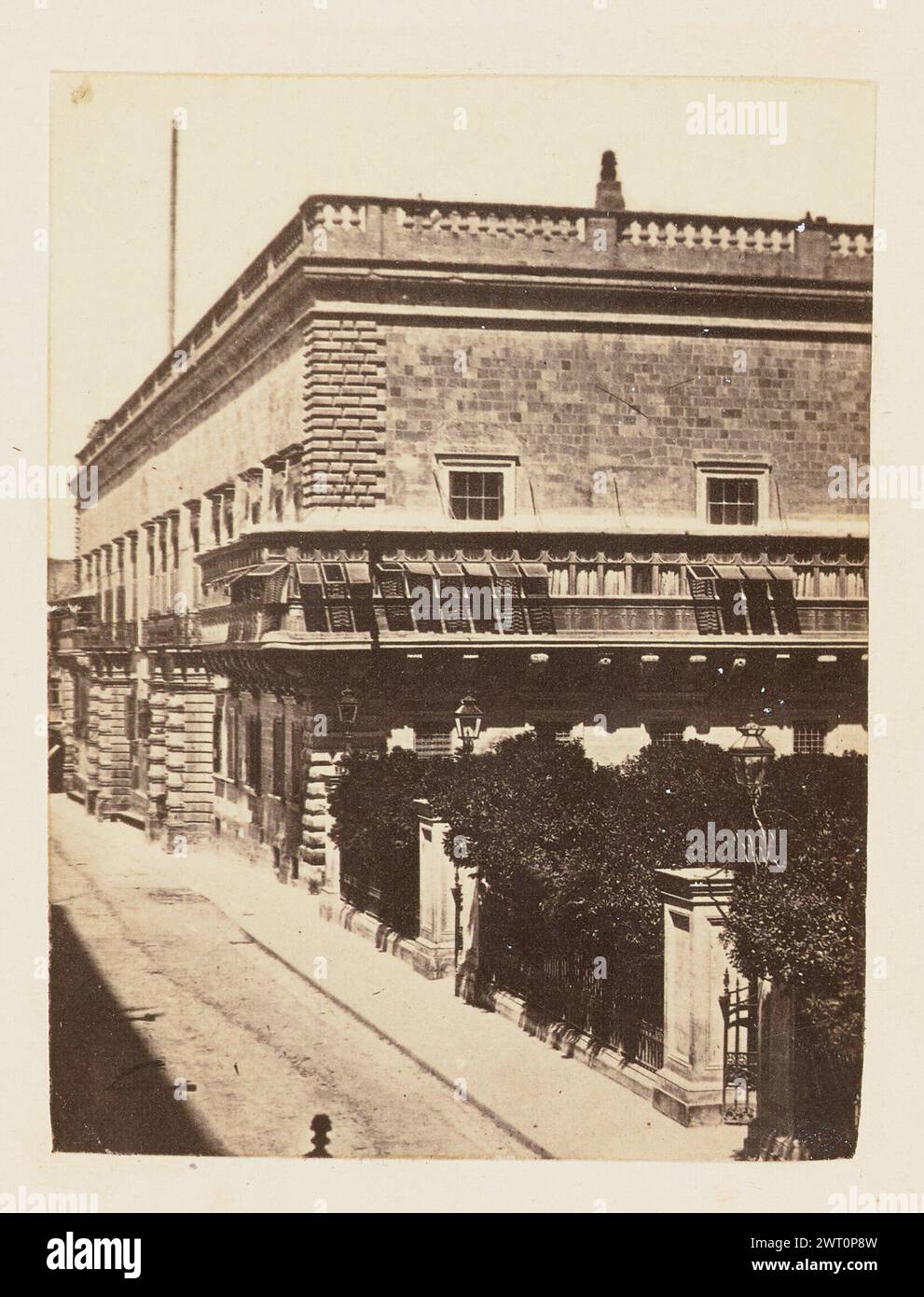 Grand Master's Palace, Valletta. Unknown, photographer about 1864 Three-quarter view of a two-story stone building with an enclosed, wooden balcony wrapped around its second-story corner and a parapet along the roof's perimeter. Also visible in the foreground is a densely grown vegetation, possibly part of a fenced-in garden. Stock Photo