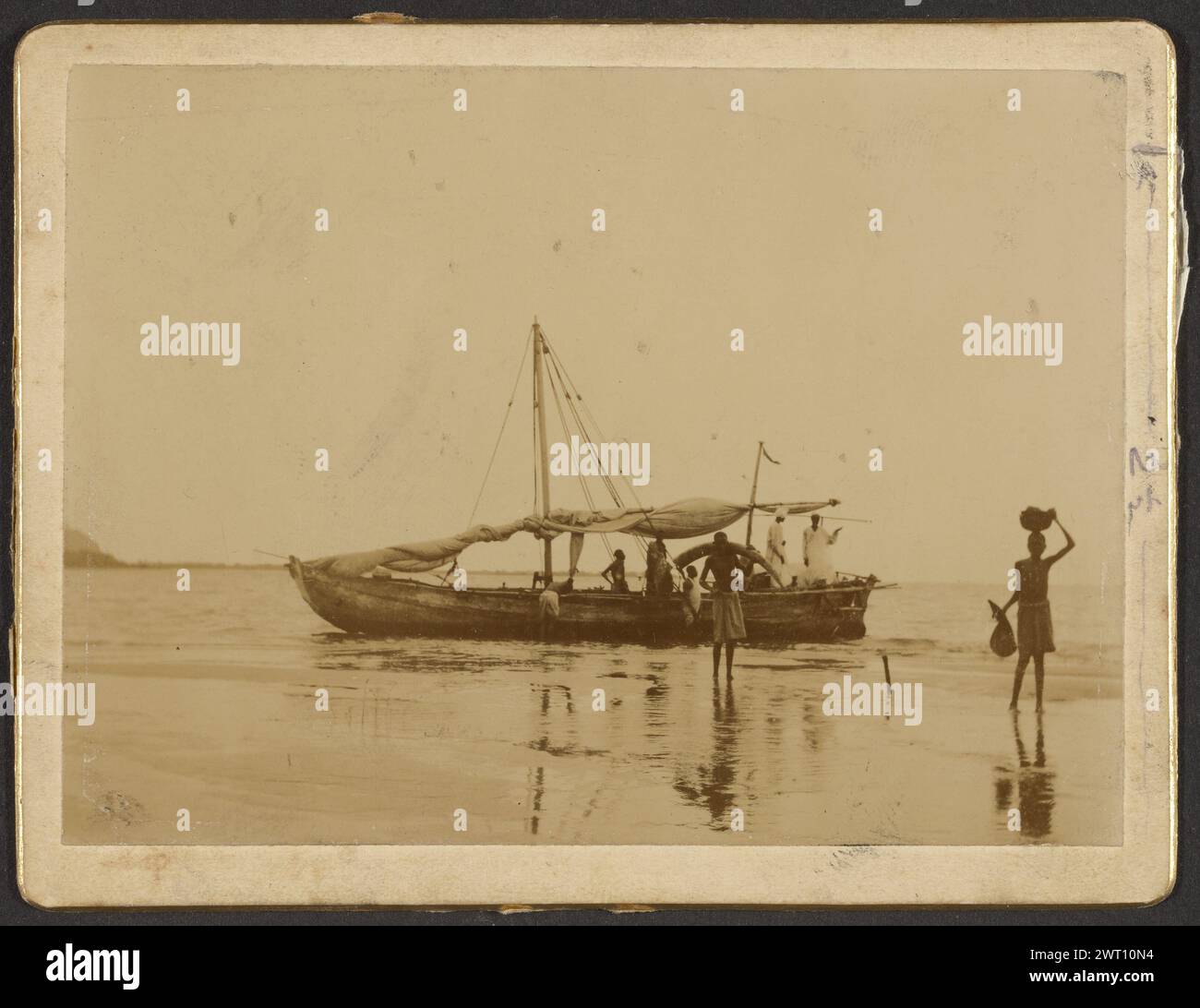 A Slave-Dhow. Unknown, photographer about 1880–1895 A long wooden boat on the shore with the sail rolled up. A figure is carrying what appears to be an elephant tusk away from the ship. Another figure is standing on the shore and is balancing a basket on their head. (Recto, mount) center right, pencil: '2 1/2'; (Recto, attached paper) center, blue pencil: '[illeg]- A slave dhow landing/ivory in the olden/days- [space] Book [pencil]'; (Verso, mount) center, pencil: 'Landing Ivory/from old slave Dhow[in black ink]/from Dhow[crossed out]/South[in black ink]/T-[illeg]-any [illeg]'; (Verso, mount) Stock Photo