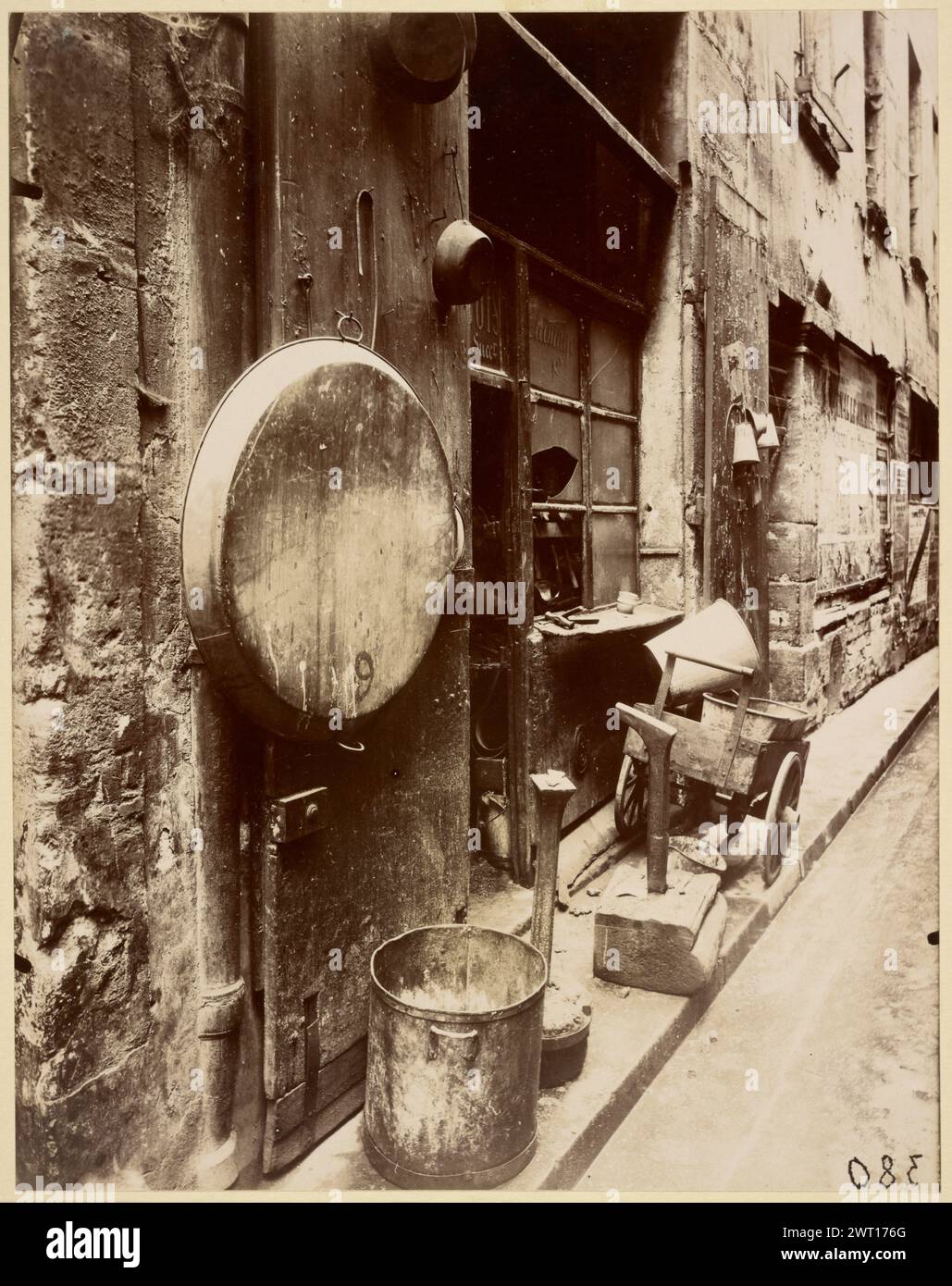 Rue de la Reynie Tin Smith's shop. Eugène Atget, photographer (French, 1857 - 1927) 1912 This bleak view documents the commercial use by the urban working class of cramped public or quasi-public spaces. In this photograph, the implements from a tinsmith's shop have spilled out onto the sidewalk. Judging by the tools on the windowsill and the scraps on the pavement, the smith worked in the street itself, traffic permitting. Teapots, various pots and pans, and a large, round basin hang next to the darkened shop door and grimy window. A barrow for picking up damaged goods and delivering mended ut Stock Photo
