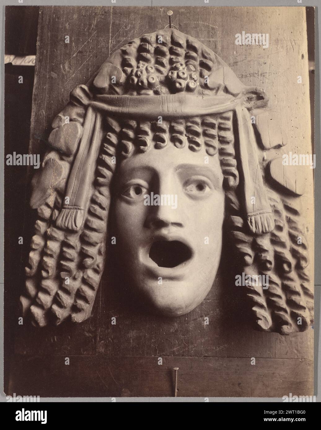 Masque antique. Eugène Atget, photographer (French, 1857 - 1927) 1923 View of a mask hanging on a nail affixed to wood. The mask's mouth is open and its eyes are wide. The mask has ringlets and is wearing a headpiece. (Verso, print) upper center, in pencil in the artist's hand: 'Masque antique'; (Verso, print) upper right, in pencil: '[illeg.] 1)' (Verso, print) upper right, in pencil in the artist's hand: '54'; (Verso, print) center, in pencil: 'S.S.'; (Verso, print) center right, in pencil in the artist's hand: '17 bis' ['bis' underlined]; (Verso, print) lower center, in pencil: 'B.A.'; (Ver Stock Photo