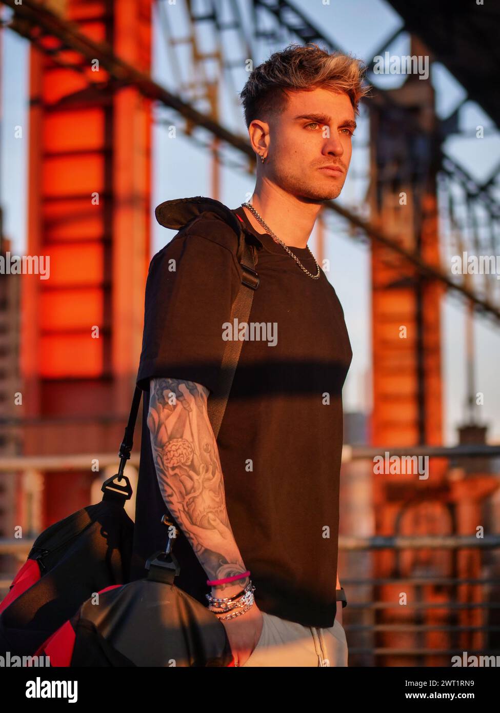 A man with a visible tattoo on his arm stands confidently in front of a modern building. He gazes ahead, creating a striking contrast between his edgy Stock Photo