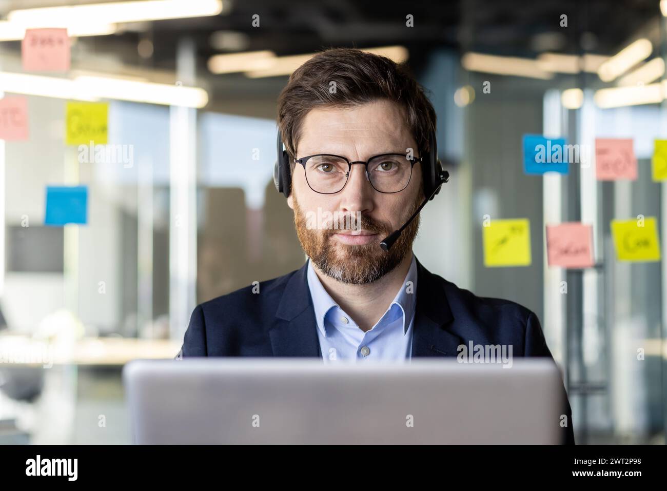 Professional male in business attire with headset using a laptop, sticky notes in background, in an office setting. Stock Photo