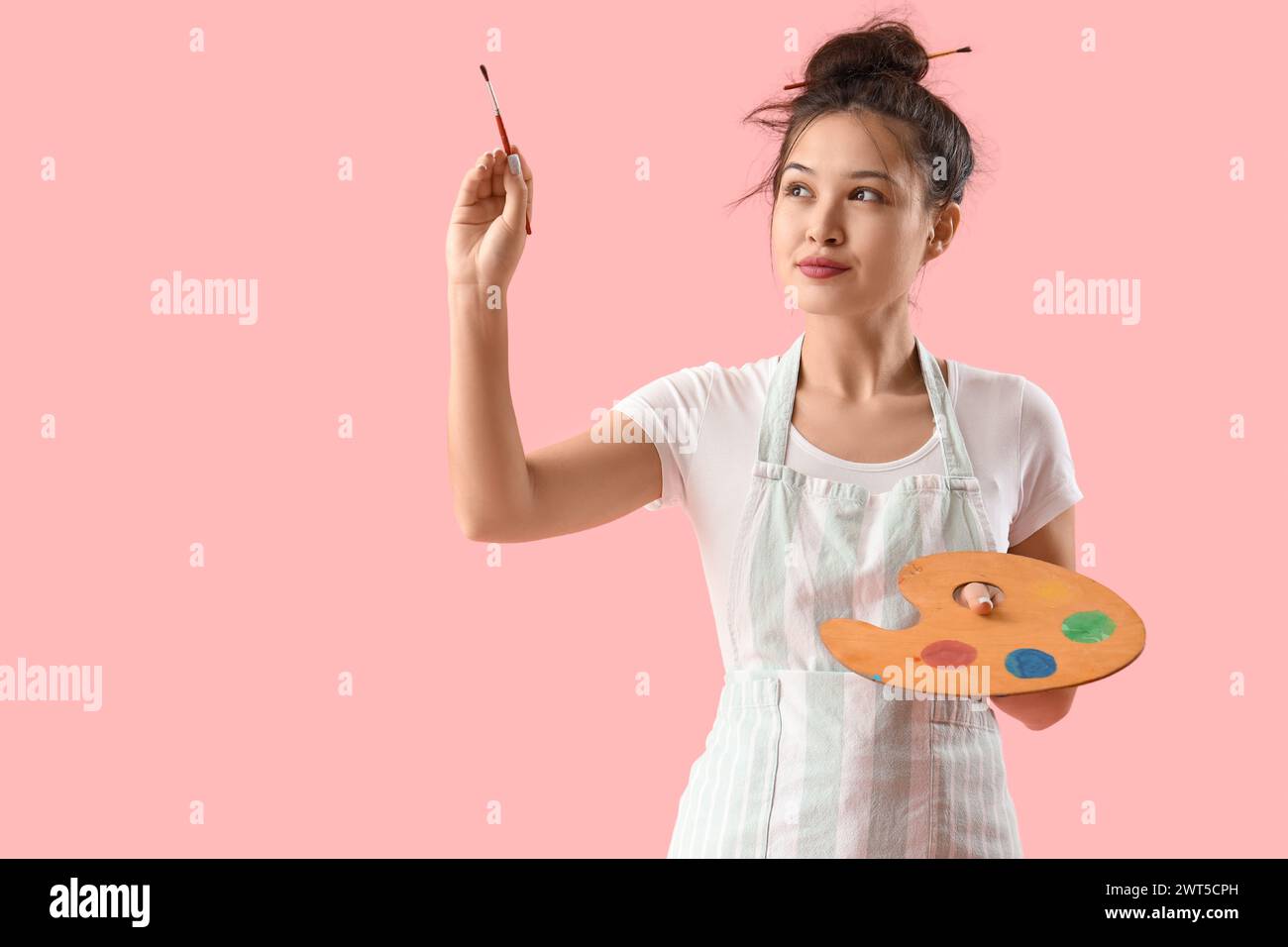 Female Asian artist with paint palette and brush on pink background ...