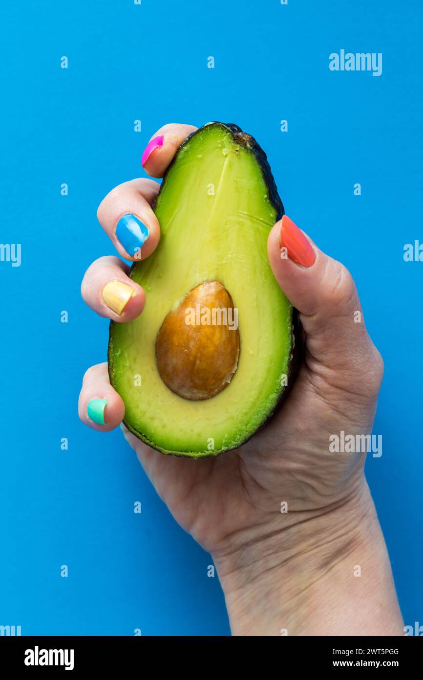 A hand with colourful nail polish holding a fresh cut avocado. Stock Photo