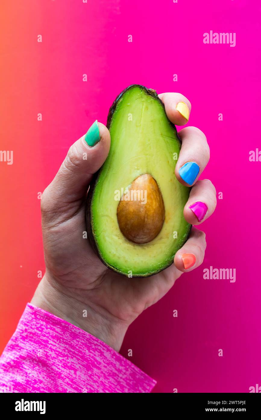 A hand with colourful nail polish holding a freshly cut avocado. Stock Photo
