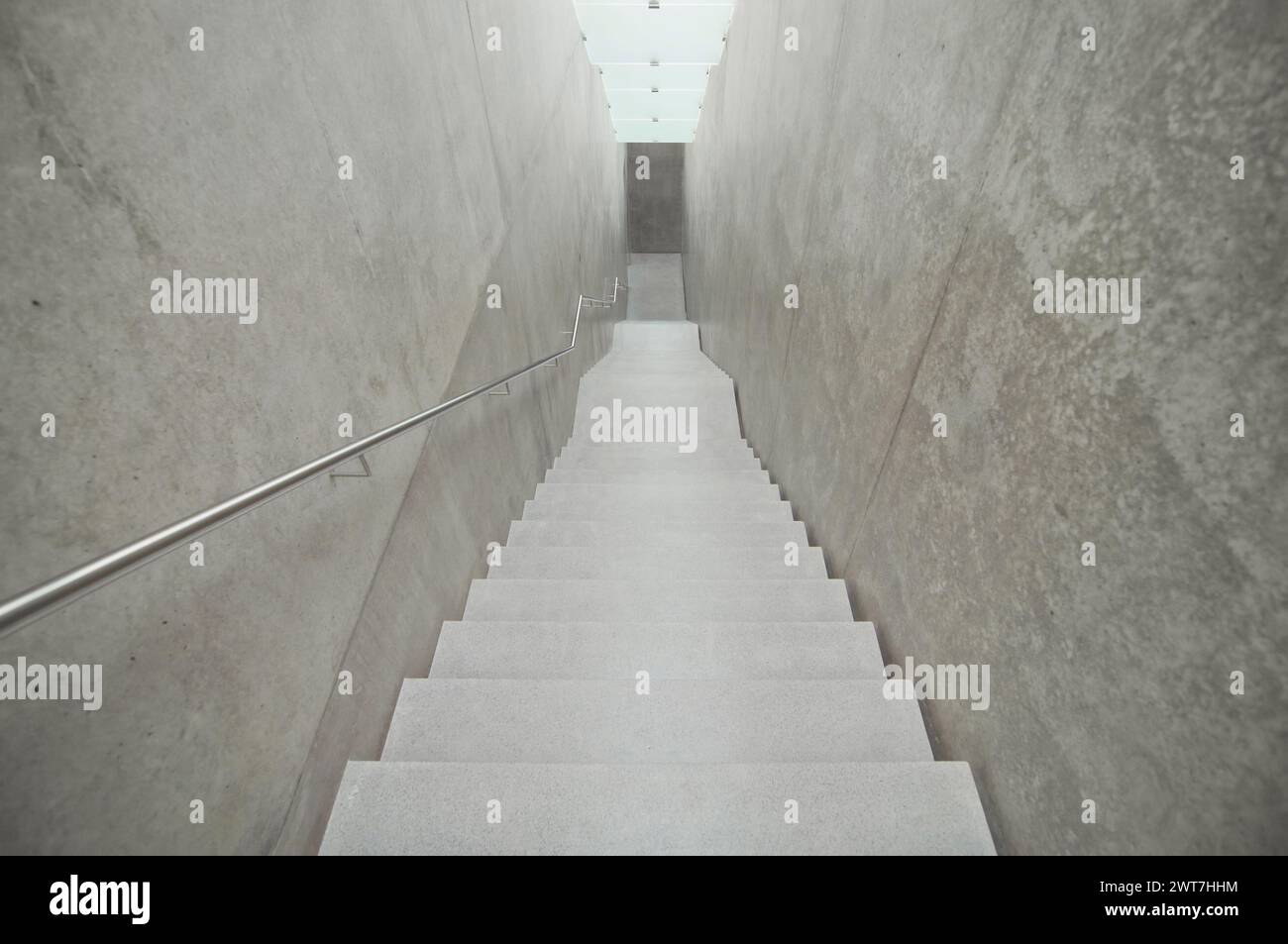 Empty narrow downwards staircase. Concrete walls and illuminated ceiling made of satin glass. Handrail on the left side. Photo in grey tones. Stock Photo