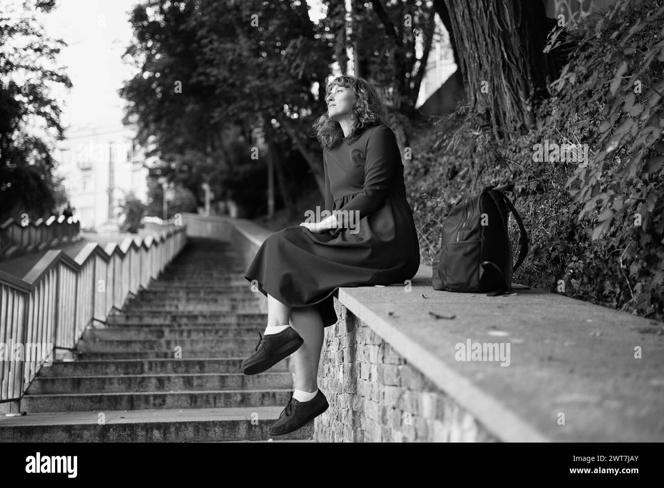 Cheerful young caucasian woman smiles with her teeth looking to side, sitting in European city on pavement. Brunette wears dress. City life concept. High quality photo Stock Photo
