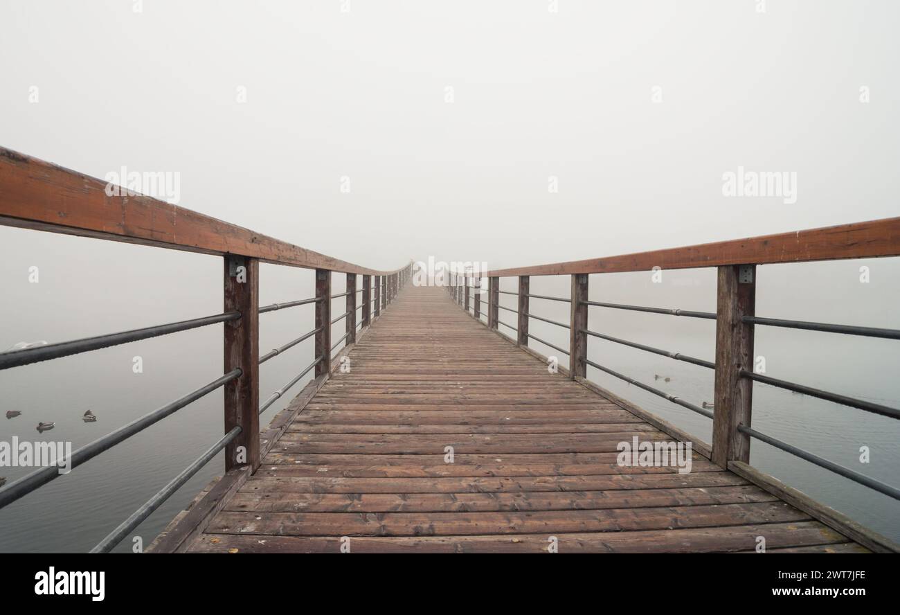 A wooden boardwalk with hand railings over a lake in the fog. Wooden walkway over still water. The walkway curves away from the viewer to the right. Stock Photo