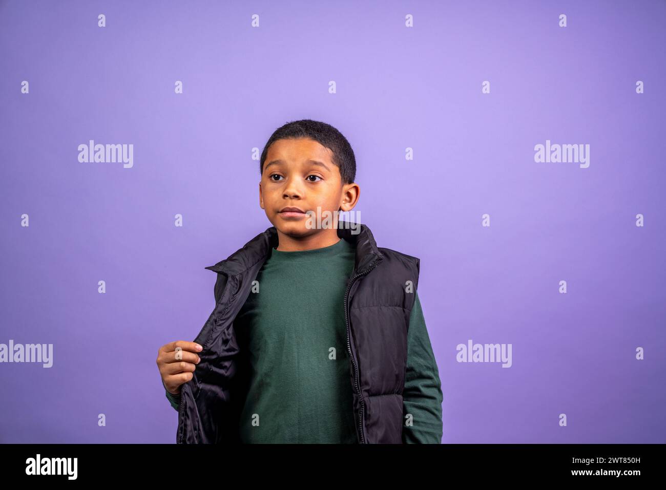 A young boy wearing a green shirt and black vest stands in front of a purple background. He looks serious and focused Stock Photo