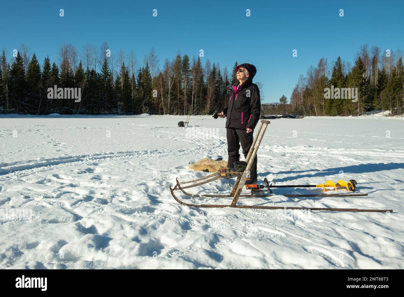 Elderly wiman stands on the ice and fishes, enjoying the spring sun and in the foreground the special Norrland winter vehicle (the kick). The picture Stock Photo