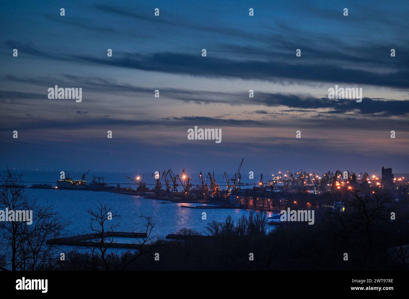 Twilight descends on busy port with cranes silhouetted against dusk sky. Maritime industrial hub transitions to night, highlighting harbor activity Stock Photo