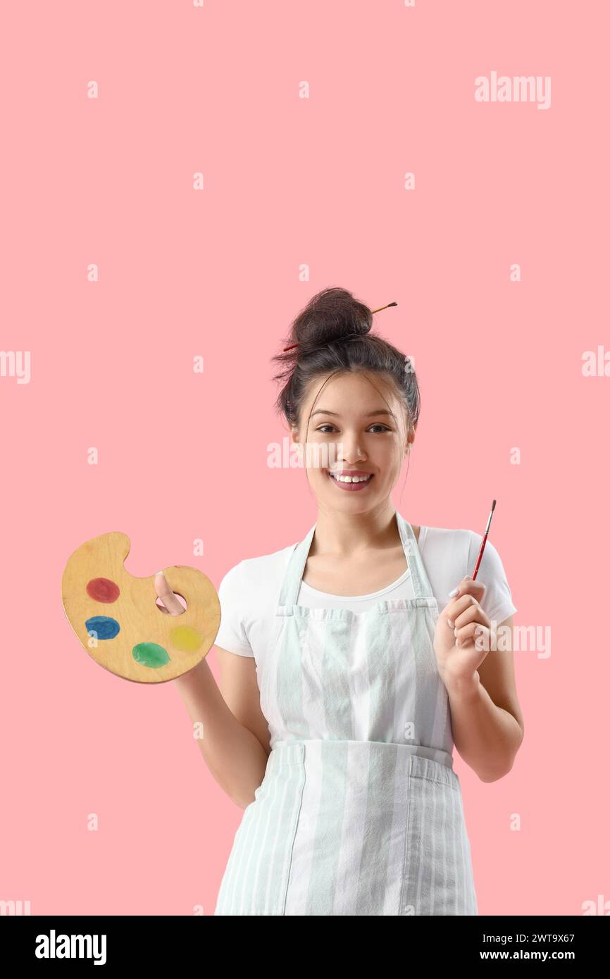 Female Asian artist with paint palette and brush on pink background ...