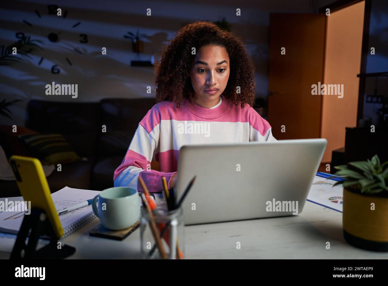 Young tired African American university woman typing on laptop sitting at desk in living room. Stock Photo