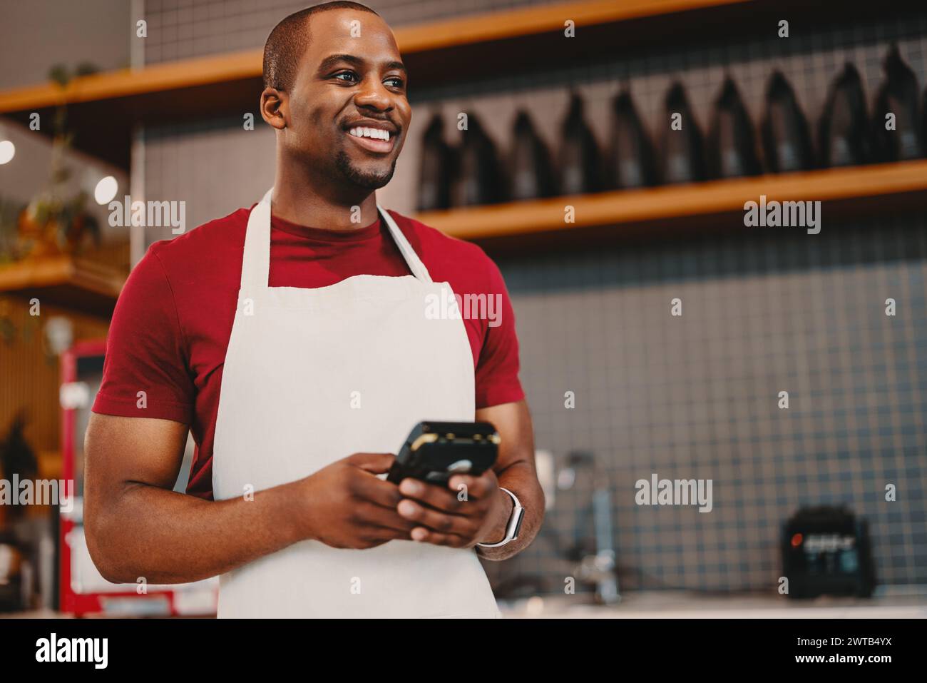 Cheerful entrepreneur holding a pos machine in a cafe, wearing a white apron. Man smiling while managing his business digitally, representing small bu Stock Photo