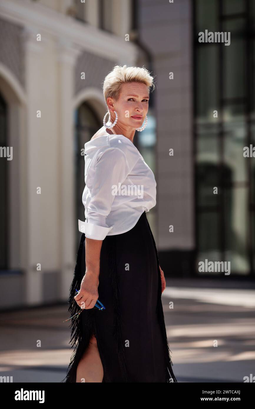 Stylish middle aged fashionista, dressed in trendy attire black skirt and a white blouse, strolls street, glancing back with elegance. Beautiful matur Stock Photo